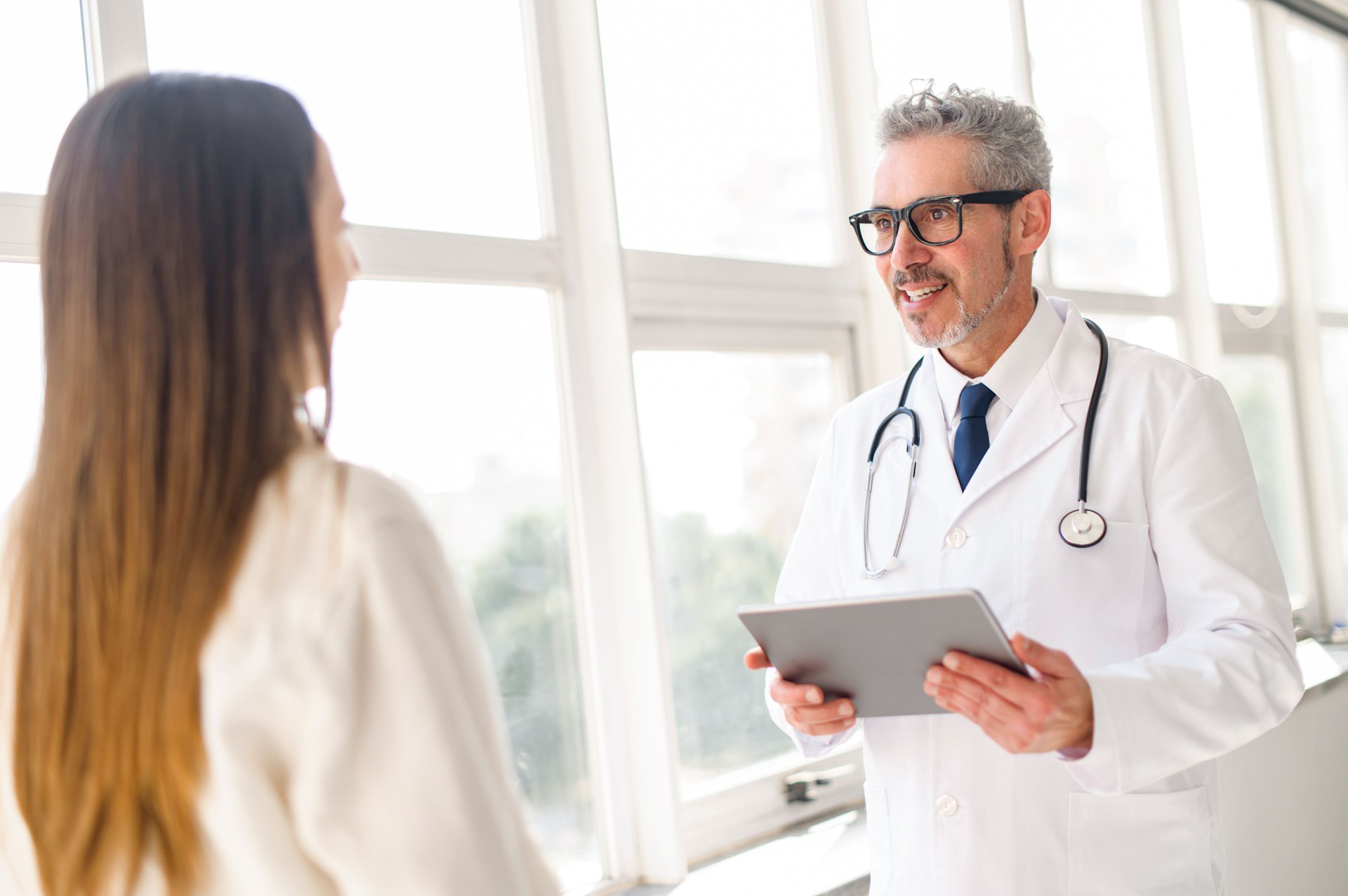 A senior doctor in a white coat is consulting with a young female patient by the window