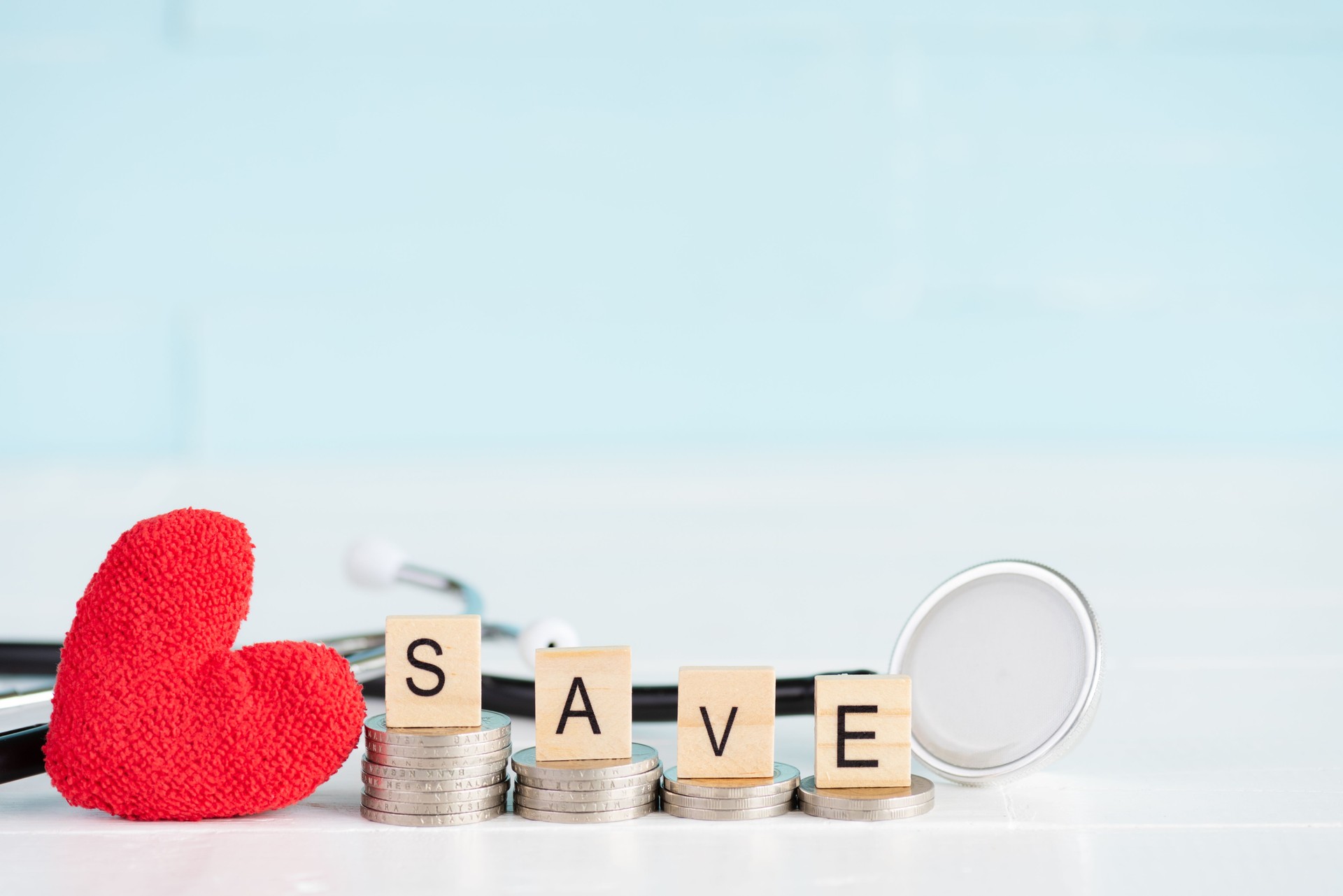 Money saving and health care concept. SAVE text on stack of coins with stethoscope and handmade red heart on white and pastel green wooden background.